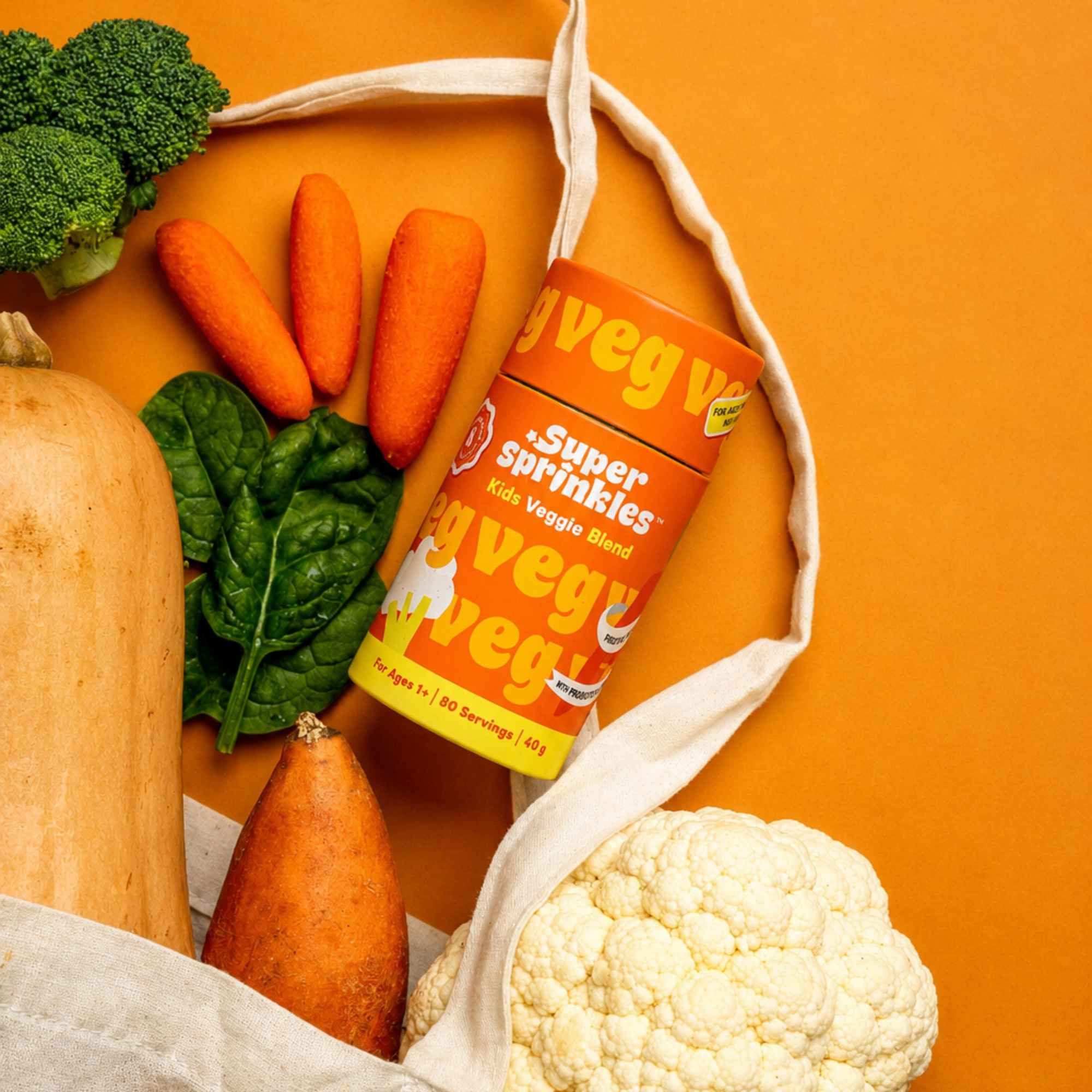 Super Sprinkles  on an orange background with fresh vegetables like broccoli, carrots, spinach, squash, sweet potato, and cauliflower arranged around it, with a cream canvas tote bag draped behind.
