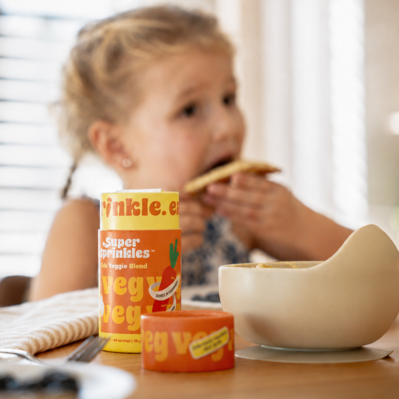A tube of Super Sprinkles Kids Veggie Blend sits on a kitchen table while a child enjoys a familiar meal in the softly blurred background, showing an easy way to add real vegetables to everyday foods for picky eaters.