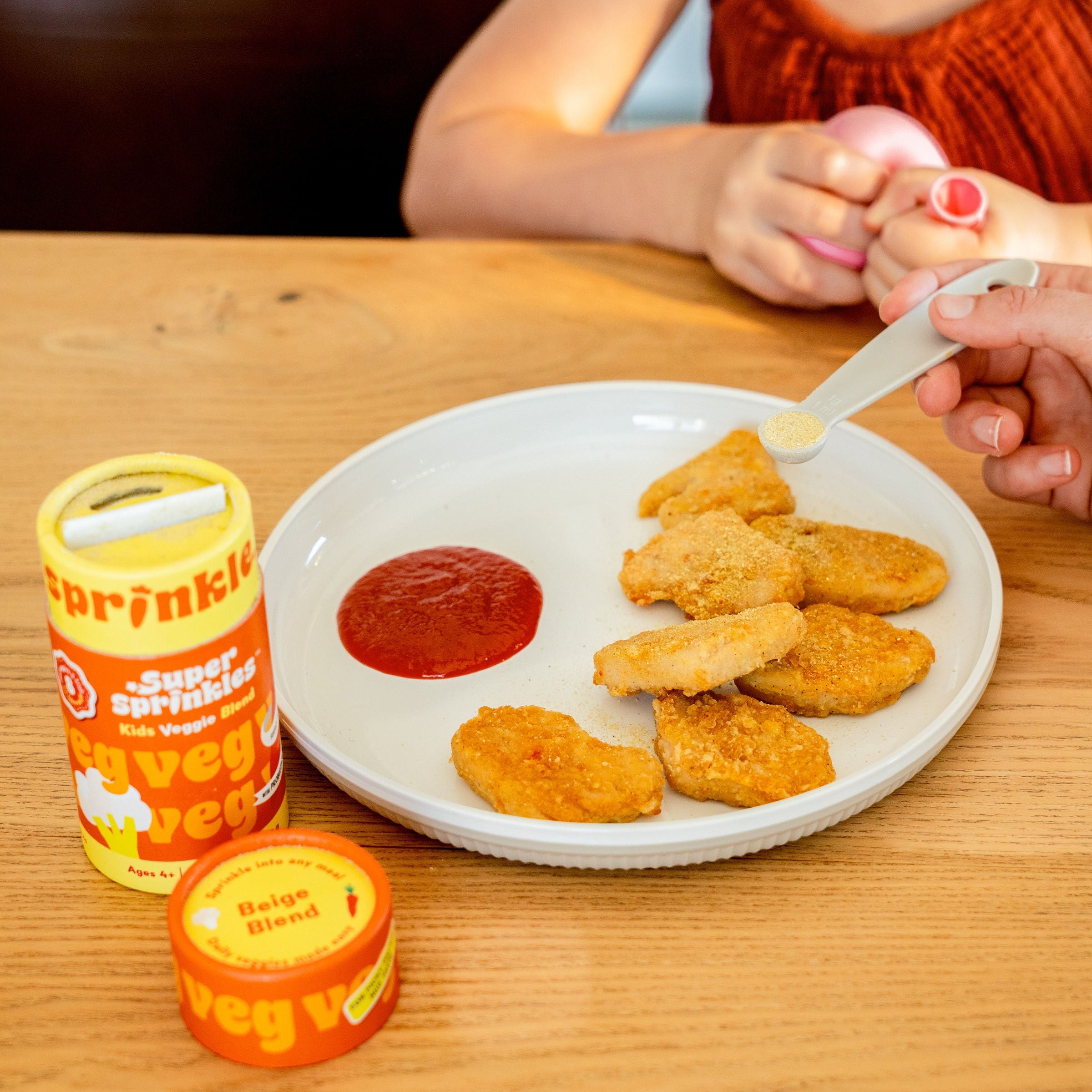 Child eating chicken nuggets with ketchup from a plate, with super sprinkles being added with a measuring spoon, on a wooden table.