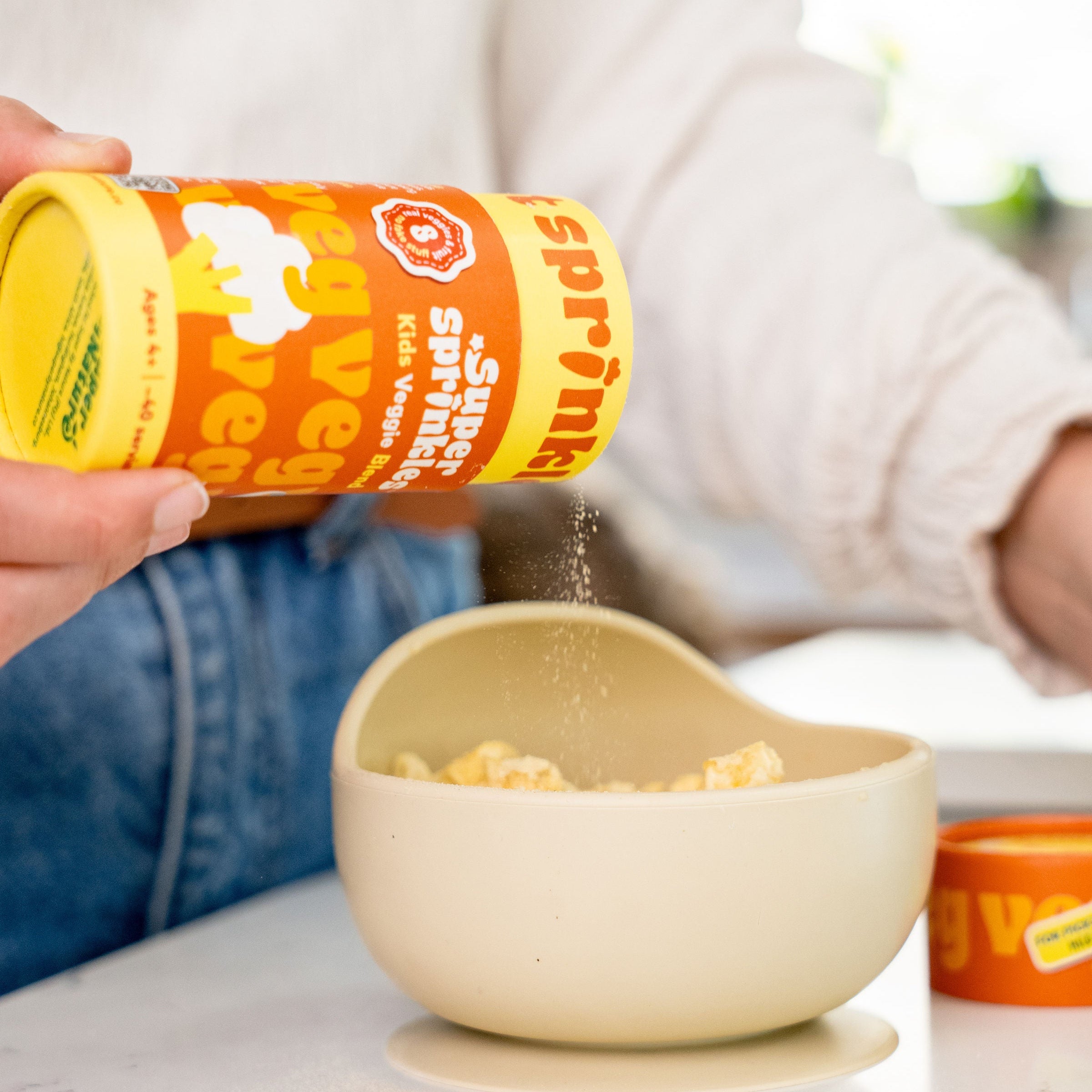 A person in a white long-sleeve top and blue jeans sprinkles super sprinkles container into a beige bowl on a white table.